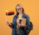 Happy young blonde lady student in casual with glasses, backpack and books holding small flag of